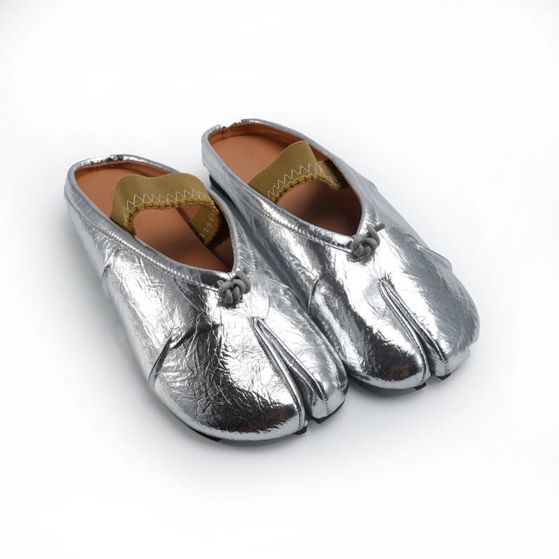 May include: A pair of silver split-toe shoes with a metallic finish. The shoes have a brown insole and a khaki-colored elastic strap. The toe area is split, and the shoes are on a white background.