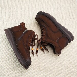 May include: A pair of brown leather ankle boots with a zipper closure and lace-up design. The boots have a rounded toe and a thick sole. The laces are adorned with wooden beads. The boots are displayed on a cream-colored surface.
