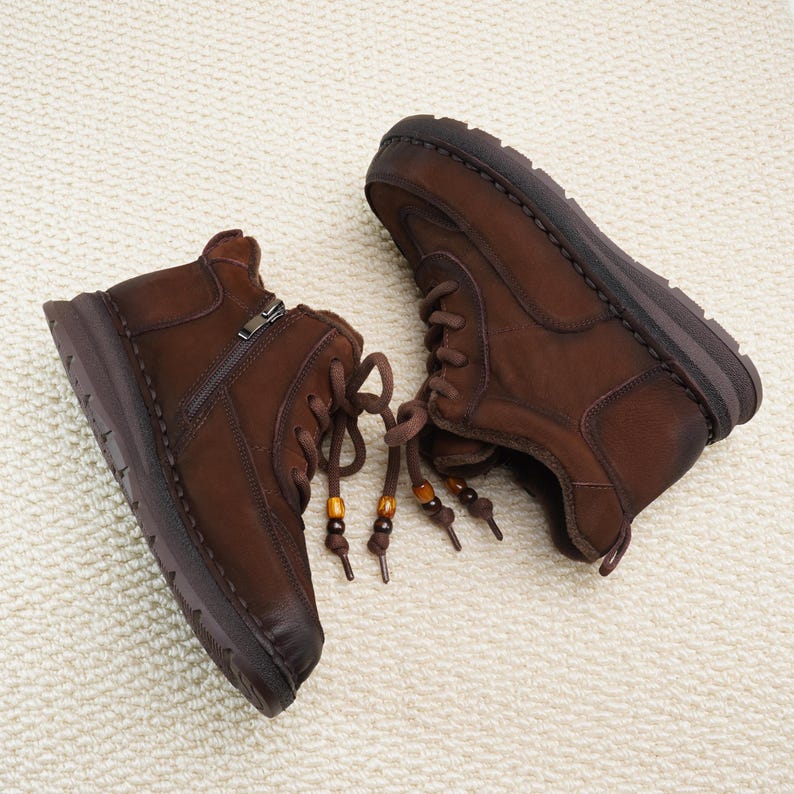 May include: A pair of brown leather ankle boots with a zipper closure and lace-up design. The boots have a rounded toe and a thick sole. The laces are adorned with wooden beads. The boots are displayed on a cream-colored surface.