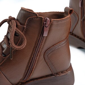 Puede incluir: Primer plano de una bota de tobillo de cuero marr&oacute;n con cremallera lateral y cordones marrones. La bota tiene una punta redondeada y un tac&oacute;n bajo. El cuero parece liso y tiene un ligero brillo. Los cordones son gruesos y anudados.