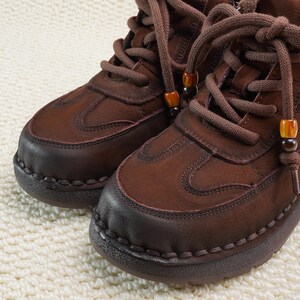 May include: Close-up of two brown leather boots with thick soles and matching laces. The boots have a rounded toe and decorative stitching around the sole. The laces are adorned with small, decorative beads. The boots are on a cream-colored, textured surface.