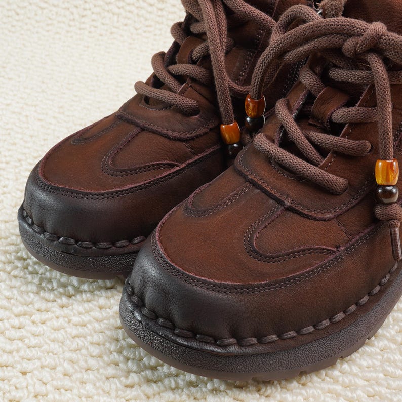 May include: Close-up of two brown leather boots with thick soles and matching laces. The boots have a rounded toe and decorative stitching around the sole. The laces are adorned with small, decorative beads. The boots are on a cream-colored, textured surface.