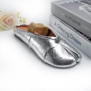 May include: Silver metallic split-toe flats with a knot detail on the vamp. The shoes have a low heel and a tan sock insert. The flats are displayed on a white surface with a stack of books and a flower in the background.
