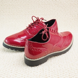 Peut inclure: Une paire de chaussures Oxford rouges brillantes avec des perforations d&eacute;coratives et des lacets rouges assortis. Les chaussures ont une semelle noire &eacute;paisse avec une bordure gris clair. Elles sont pos&eacute;es sur une surface textur&eacute;e blanc cass&eacute;.