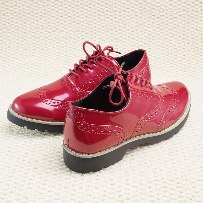 Peut inclure: Une paire de chaussures Oxford rouges brillantes avec des perforations d&eacute;coratives et des lacets rouges assortis. Les chaussures ont une semelle noire &eacute;paisse avec une bordure gris clair. Elles sont pos&eacute;es sur une surface textur&eacute;e blanc cass&eacute;.