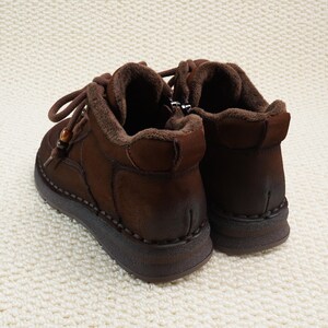May include: A pair of brown leather ankle boots with a warm, fuzzy interior. The boots have a lace-up design with a wooden bead detail and a zipper on the side. The soles are thick and dark brown, and the boots are set against a white, textured background.