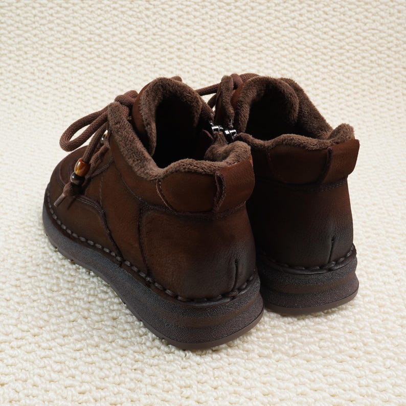 May include: A pair of brown leather ankle boots with a warm, fuzzy interior. The boots have a lace-up design with a wooden bead detail and a zipper on the side. The soles are thick and dark brown, and the boots are set against a white, textured background.