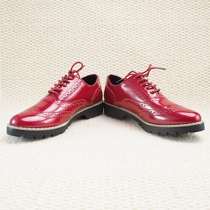 Peut inclure: Une paire de chaussures Oxford en cuir verni rouge avec un motif ajour&eacute;. Les chaussures ont une finition brillante, des lacets assortis et une semelle noire &eacute;paisse avec une bordure de couleur claire. Elles sont pos&eacute;es sur une surface textur&eacute;e blanc cass&eacute;.