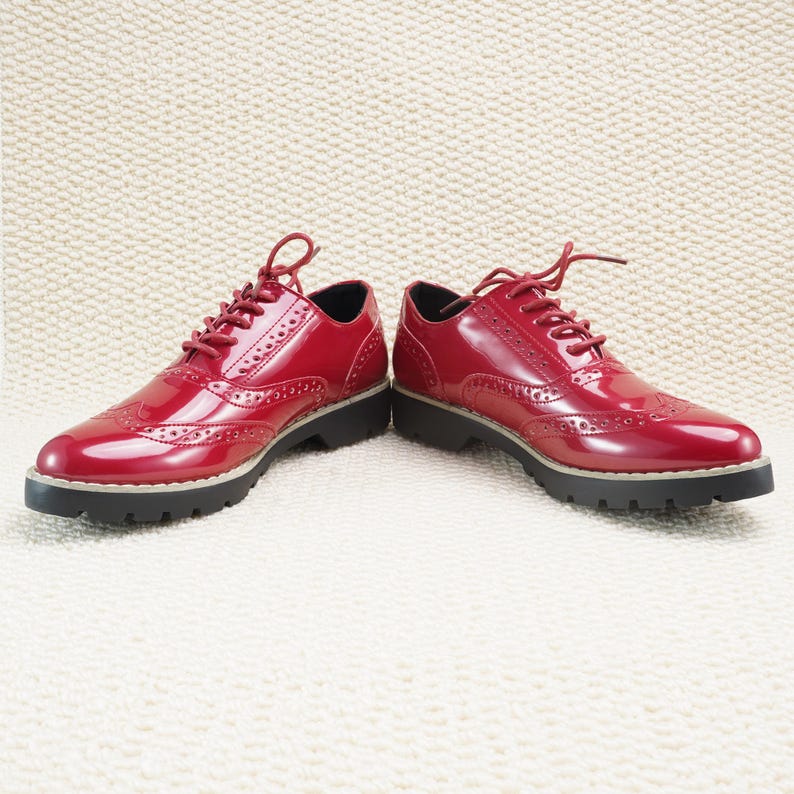 Peut inclure: Une paire de chaussures Oxford en cuir verni rouge avec un motif ajour&eacute;. Les chaussures ont une finition brillante, des lacets assortis et une semelle noire &eacute;paisse avec une bordure de couleur claire. Elles sont pos&eacute;es sur une surface textur&eacute;e blanc cass&eacute;.