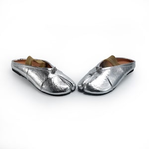 May include: A pair of silver metallic split-toe slip-on shoes. The shoes have a rounded toe and a small bow detail at the top. The interior is lined with a tan material. The shoes are designed to be worn without a back.