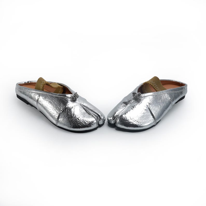 May include: A pair of silver metallic split-toe slip-on shoes. The shoes have a rounded toe and a small bow detail at the top. The interior is lined with a tan material. The shoes are designed to be worn without a back.
