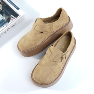 Pode incluir: Um par de sapatos de camurça bege com sola castanha grossa. Cada sapato tem uma tira com fivela dourada e bico arredondado. Os sapatos são exibidos em uma superfície branca.