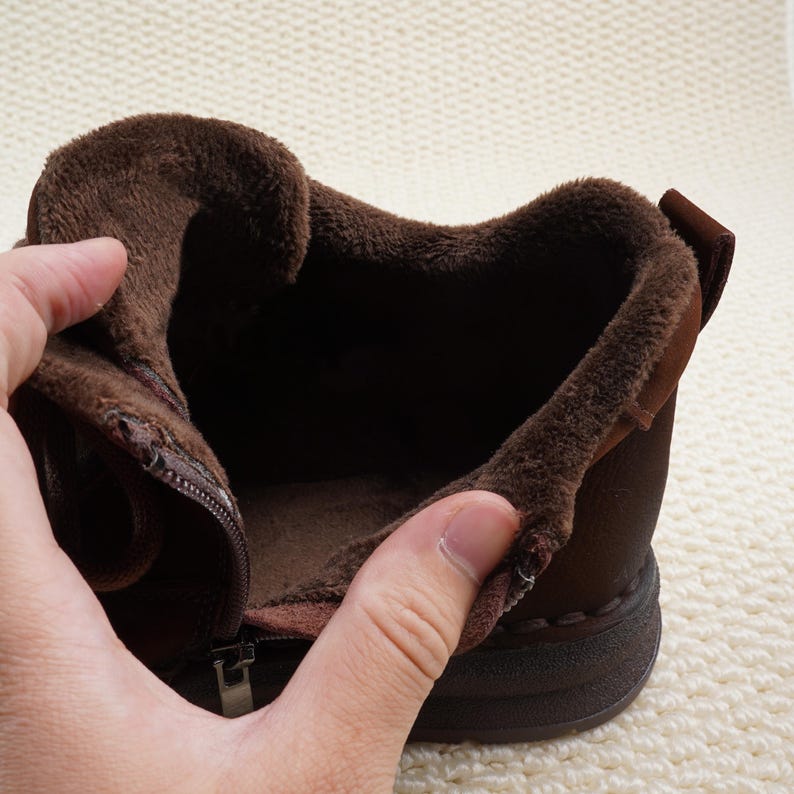 May include: A brown boot with a zipper, featuring a plush, dark brown interior lining. The boot's exterior is a darker shade of brown, and the sole is a solid, darker brown. The boot is designed for warmth and comfort.