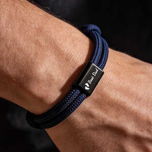 Könnte beinhalten: Marineblaues Seilarmband mit schwarzem Verschluss. Der Verschluss ist mit den Worten "Best Dad" und einem kleinen Fußabdruck-Symbol graviert. Das Armband wird am Handgelenk getragen, vor dunklem Hintergrund.