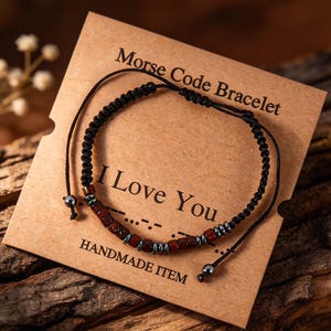 May include: A black cord bracelet featuring small dark red and metallic grey beads arranged in a Morse code pattern. The bracelet is displayed on a tan card printed with the text Morse Code Bracelet, I Love You, and HANDMADE ITEM.