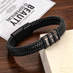May include: Black braided leather bracelet with a magnetic clasp. The bracelet features three metal beads engraved with the names "Hazel", "Luke", and "Leo". The text "BEAUTY IS SUPERFICIAL" is visible in the background.