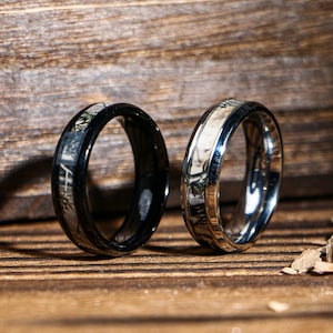 Puede incluir: Dos anillos con diseño de vetas de madera. Uno es negro con incrustaciones de vetas de madera, y el otro es plateado con un patrón similar. Los anillos se exhiben sobre una superficie de madera.