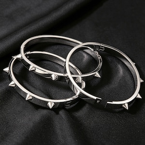 May include: Three silver spiked bracelets are shown against a dark backdrop. The bracelets are circular, with evenly spaced, pointed spikes. One bracelet is partially unclasped, showing the clasp mechanism.