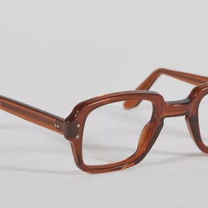 Könnte beinhalten: Braun getönte Brille mit rechteckigem Rahmen. Die Brille hat ein klassisches Design mit leicht abgerundeter Form und sichtbaren Scharnieren. Die Gläser sind klar und die Bügel haben eine passende braune Farbe.
