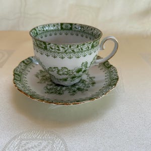 Può includere: Servizio da tè bianco con motivo floreale verde e bordi dorati. La tazza ha un manico curvo e il piattino è rotondo. Il set è appoggiato su una tovaglia bianca.