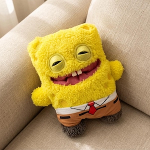May include: A yellow plush character with a large, smiling mouth and closed eyes. The character wears brown trousers, a white shirt, and a red tie. The soft toy is on a beige sofa.