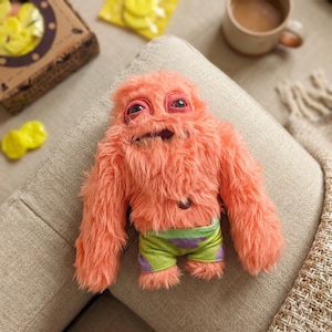 May include: A soft, peach-coloured monster doll with a shaggy texture. It has large, expressive eyes and wears green shorts with purple accents. The doll is posed on a neutral-toned surface.