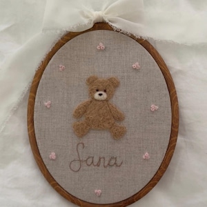 May include: An oval embroidery hoop with a brown teddy bear and the name "Sana" embroidered on linen fabric. Pink floral details and a wooden frame. A white ribbon bow is attached at the top.