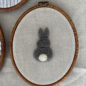 May include: A grey felted bunny with a white tail is centered on a light grey fabric, set within a wooden embroidery hoop. The hoop is part of a set of three, with the other two featuring different fabric patterns.
