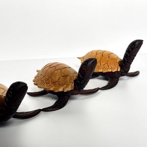 Puede incluir: Tres figuras de tortugas de madera. Las tortugas tienen cabezas, patas y colas de color marrón oscuro, con caparazones texturizados de color marrón claro. Las tortugas están dispuestas en fila, con la más cercana enfocada.