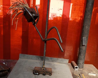 May include: A welded metal sculpture of a figure with a square head and wire hair. The figure is made of rebar and sits on a wooden base. The background is a red-orange wall.