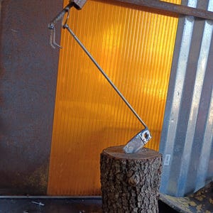 May include: A handcrafted metal garden hoe with a wooden handle and a rectangular blade, resting on a tree stump. The hoe is made of silver metal and has a rustic appearance. The background features corrugated metal and a yellow-orange translucent panel.