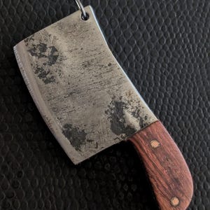 May include: A miniature cleaver pendant with a distressed metal blade and a dark wood handle. The small cleaver has a silver-colored loop for attaching to a chain or keychain. The handle is secured with two small rivets.