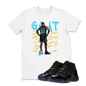 May include: White t-shirt featuring a basketball player graphic and the word "GOAT" in turquoise. Black and turquoise basketball trainers are in the foreground. The shirt design includes the number 23 and other text.