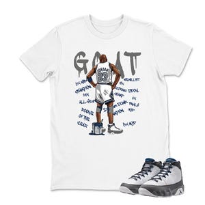 May include: White t-shirt with a graphic of a basketball player and the word "GOAT" in gray. The shirt features text and a pair of gray and white high-top sneakers. The text includes "6X NEW CHAMPION", "ROOKIE OF THE YEAR", and "JORDAN 23".