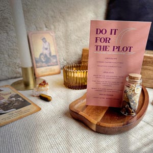 May include: A collection of spiritual items including a lit candle, tarot cards, and a small glass jar filled with crystals. A pink card with the text "DO IT FOR THE PLOT" is displayed on a wooden tray. The scene is set on a textured, neutral-coloured surface.