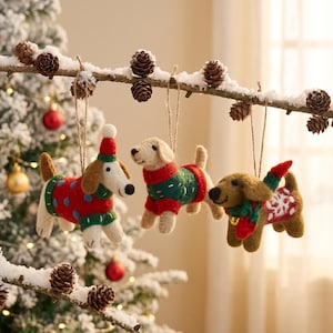 Può includere: Tre decorazioni a forma di cane in feltro appese a un ramo innevato con pigne. I cani indossano maglioni e cappelli festivi rossi, verdi e bianchi, perfetti per l'albero di Natale.