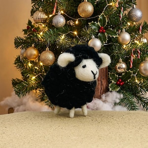May include: A needle-felted sheep figurine with a black wool body and white wool face, ears, and legs. The sheep has small black eyes and a stitched black nose, standing on a textured surface in front of a decorated Christmas tree.