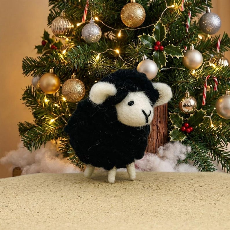 May include: A needle-felted sheep figurine with a black wool body and white wool face, ears, and legs. The sheep has small black eyes and a stitched black nose, standing on a textured surface in front of a decorated Christmas tree.