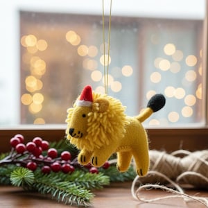 May include: A yellow felt lion Christmas ornament with a red and white Santa hat. The lion has a black tail and is hanging from a gold string. The background includes a window with blurred lights and holiday greenery.