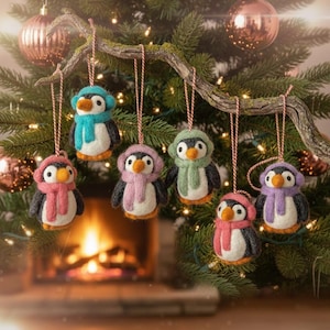 Puede incluir: Adornos navideños de pingüinos hechos a mano colgando de una rama de árbol de Navidad. Cada pingüino lleva un gorro y una bufanda en varios colores, incluyendo azul, rosa, verde y morado. Los adornos están suspendidos por hilo rojo y blanco.