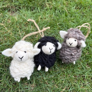 May include: Three small sheep ornaments made of textured yarn. One sheep is white, one is black, and one is brown. Each sheep has a smooth felt face with a stitched nose and mouth, small ears, and a jute twine loop attached to the top for hanging.