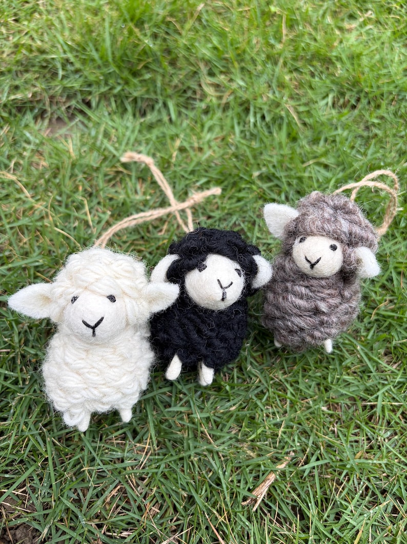 May include: Three small sheep ornaments made of textured yarn. One sheep is white, one is black, and one is brown. Each sheep has a smooth felt face with a stitched nose and mouth, small ears, and a jute twine loop attached to the top for hanging.