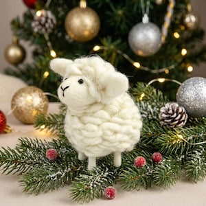 May include: A small, cream-colored needle-felted sheep figurine with a textured, woolly body and a smooth face. The sheep has two small black eyes and a stitched nose, standing upright on four short legs amidst green pine branches.