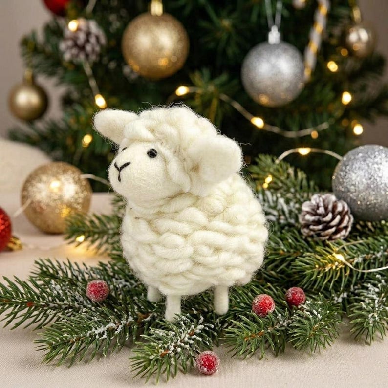 May include: A small, cream-colored needle-felted sheep figurine with a textured, woolly body and a smooth face. The sheep has two small black eyes and a stitched nose, standing upright on four short legs amidst green pine branches.
