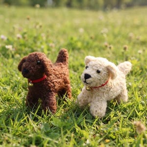 May include: Two small crocheted dog figurines, one dark brown and one cream-colored. Each dog features a textured yarn body, black bead eyes, a black stitched nose, and a thin red collar adorned with a small, round, metallic gold-colored bell.