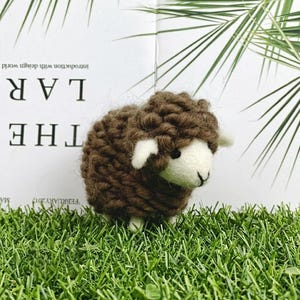 May include: A small, handcrafted sheep figurine featuring a dark brown, chunky knit wool body and a smooth, off-white felted face. The sheep has small, pointed ears, a simple stitched nose and mouth, and dark eyes, standing on four short legs.