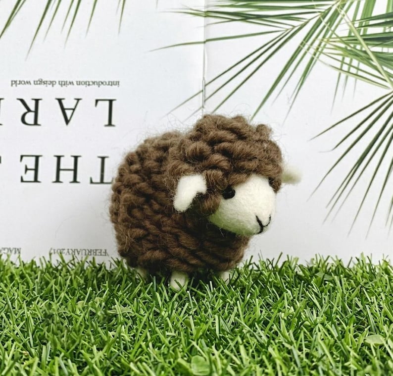 May include: A small, handcrafted sheep figurine featuring a dark brown, chunky knit wool body and a smooth, off-white felted face. The sheep has small, pointed ears, a simple stitched nose and mouth, and dark eyes, standing on four short legs.