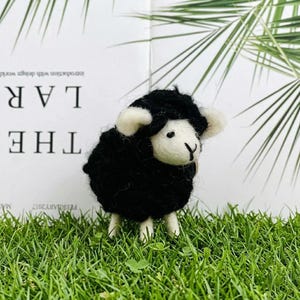 May include: A small needle-felted sheep figurine with a textured black wool body and a smooth cream-colored face. The sheep has small cream-colored ears, two tiny black eyes, and a stitched black nose. It stands on four short cream-colored legs.