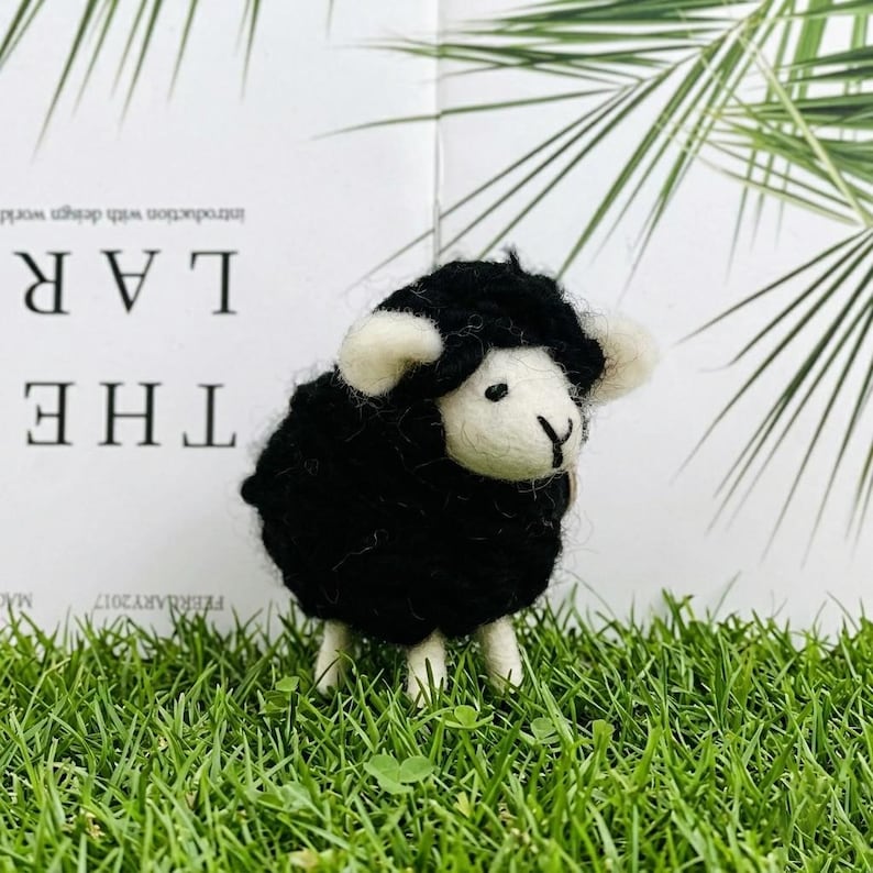 May include: A small needle-felted sheep figurine with a textured black wool body and a smooth cream-colored face. The sheep has small cream-colored ears, two tiny black eyes, and a stitched black nose. It stands on four short cream-colored legs.