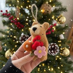 May include: A handmade brown felt bear Christmas ornament holding a bouquet of felt flowers. The flowers are red, pink, and orange with a yellow center. The ornament has a string for hanging and is set against a Christmas tree background.